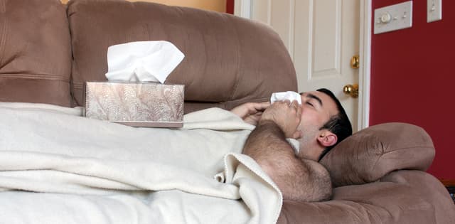 man lying on couch under a blanket sneezing into a tissue